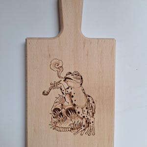 Può includere: Tagliere in legno con manico, con un disegno pirografato di una rana che fuma la pipa seduta su un teschio. La rana ha macchie nere e il teschio ha ombreggiature dettagliate. Il tagliere è in legno chiaro.