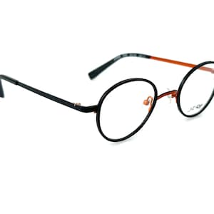 May include: Round eyeglasses with black frames and orange accents. The lenses are clear, and the temples are thin and black. The glasses have a modern, minimalist design, suitable for everyday wear. The brand name is visible on the lens.