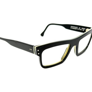 Puede incluir: Gafas rectangulares negras con una franja blanca a lo largo del borde del marco. Las gafas tienen una forma ligeramente inclinada.