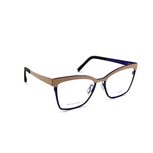 Op de afbeelding: Cat-eye bril met een goudkleurig montuur en donkerblauwe accenten. De bril heeft een strak, modern design met een dun montuur en een subtiele curve aan de bovenkant van de glazen. De pootjes zijn zwart.