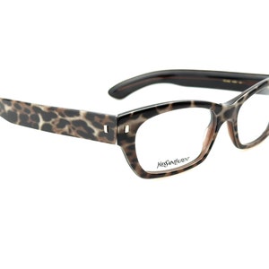 May include: Leopard print eyeglasses with brown frames and clear lenses. The brand name "Yves Saint Laurent" is printed on the right lens.