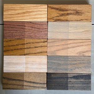 May include: Eight wood samples with different stains. Each sample is divided into two sections, one with a lighter stain and the other with a darker stain. The stains range from light brown to dark brown and black.