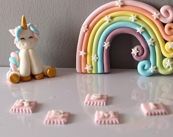 May include: A white unicorn figurine with a rainbow and stars cake topper. There are also five pink fondant squares with white numbers on them.