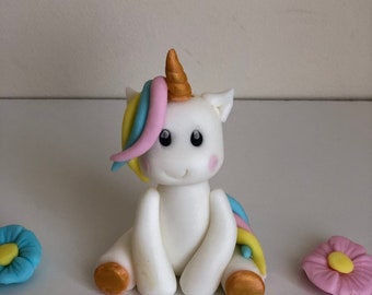 May include: A white unicorn cake topper with a rainbow mane and tail, gold hooves, and black eyes. The unicorn is sitting on a white surface with pink, yellow, and blue flowers and a pink number 4.