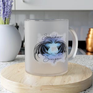 Może przedstawiać: Mug ze szkła mrożonego z uchwytem. Kubek przedstawia wzór dwóch czarnych skrzydeł smoka na niebiesko-fioletowym tle chmur. Napis "Shadow Singer" jest napisany kursywą.
