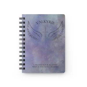 Puede incluir: Una libreta espiral de color morado y azul con un diseño de galaxia. La libreta tiene una ilustración en blanco y negro de una espada con alas y el texto "VALKYRIE" sobre la espada. El texto "I AM THE ROCK AGAINST WHICH THE SURF CRASHES" está debajo de la ilustración.