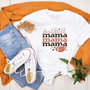 May include: White t-shirt with the text "football mama mama mama" in brown and pink lettering. A brown football with white laces is centered below the text.