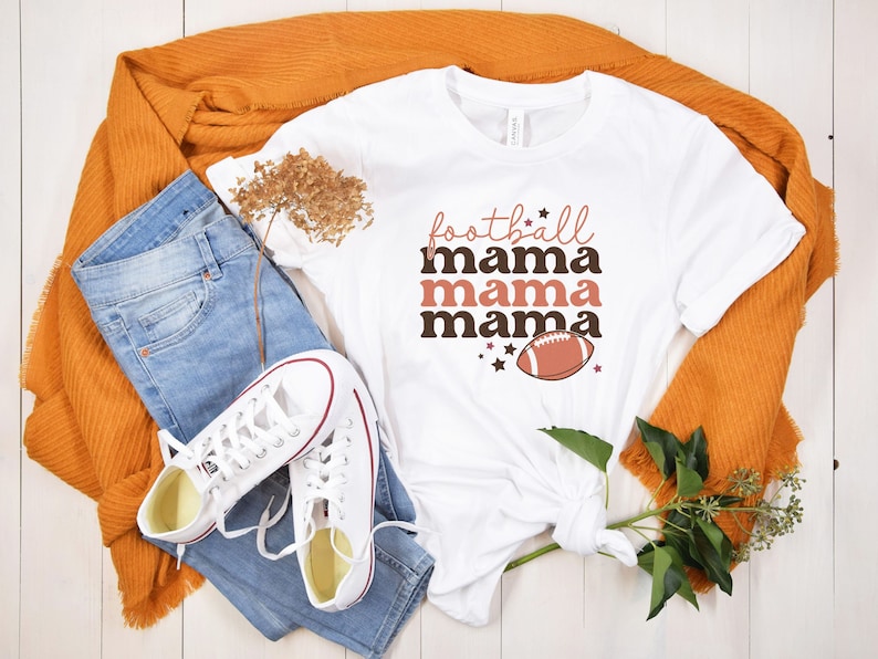 May include: White t-shirt with the text "football mama mama mama" in brown and pink lettering. A brown football with white laces is centered below the text.
