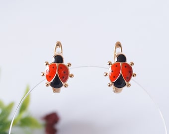 May include: A pair of gold-toned ladybug earrings with red and black enamel. The earrings are on a white background.
