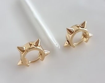 May include: A pair of gold-colored hoop earrings. Each earring features a design with three pointed spikes. The earrings are displayed on a white surface, with a soft, diffused light reflecting off the metal.