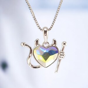 Puede incluir: Un collar de plata con un colgante en forma de corazón con una cola y cuernos de diablo. El corazón está hecho de cristal iridiscente.