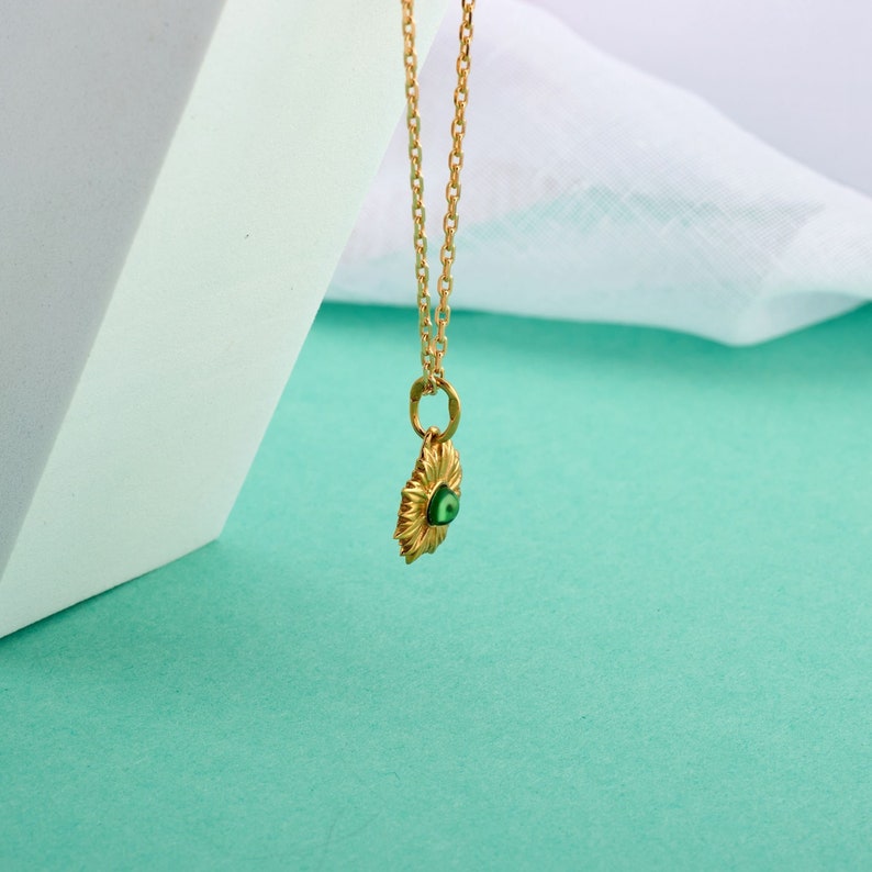 Puede incluir: Un collar de cadena dorada con un colgante que presenta un amuleto de ojo turco verde. El colgante tiene forma de sol con un dise&ntilde;o detallado.