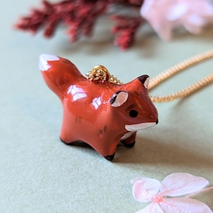 Puede incluir: Un colgante de cerámica roja en forma de zorro en una cadena de oro. El zorro está mirando hacia el lado derecho de la imagen y tiene la punta de la cola blanca.