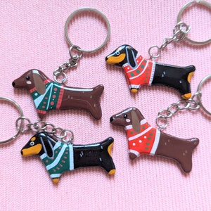 Peut inclure: Quatre porte-clés en forme de dachshund avec des pulls de couleurs différentes. Les pulls sont rouges, verts et blancs avec un motif de pois rouges et blancs. Les porte-clés sont en métal et ont une finition argentée.