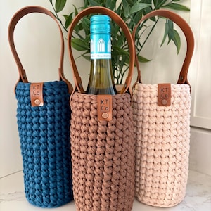 May include: Three cylindrical bottle holders made of crocheted fabric in navy blue, brown, and light pink. Each holder features a brown faux leather handle attached with metal rivets and a small brown leather tag embossed with the text KT Co.