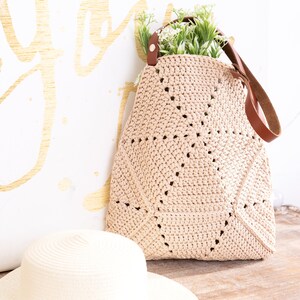 Op de afbeelding: Een beige gehaakte tote bag met bruine leren hengsels. De tas ligt op een houten oppervlak naast een beige strohoed.