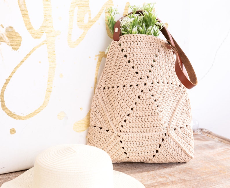 Op de afbeelding: Een beige gehaakte tote bag met bruine leren hengsels. De tas ligt op een houten oppervlak naast een beige strohoed.