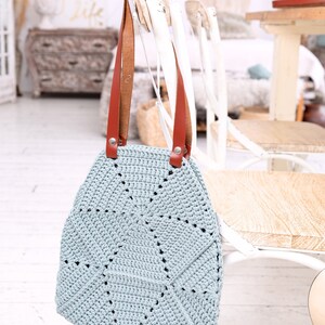 Op de afbeelding: Een lichtblauwe gehaakte tote bag met bruine leren hengsels. De tas hangt aan een witte stoel.