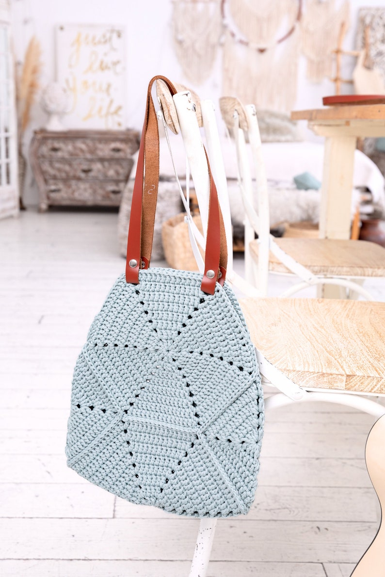 Op de afbeelding: Een lichtblauwe gehaakte tote bag met bruine leren hengsels. De tas hangt aan een witte stoel.