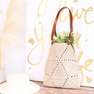 Op de afbeelding: Een beige gehaakte tote bag met bruine leren hengsels. De tas staat op een houten oppervlak voor een wit canvas met gouden letters die "I love you life" zeggen.