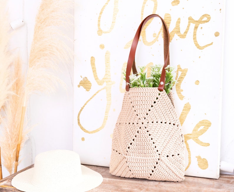 Op de afbeelding: Een beige gehaakte tote bag met bruine leren hengsels. De tas staat op een houten oppervlak voor een wit canvas met gouden letters die "I love you life" zeggen.
