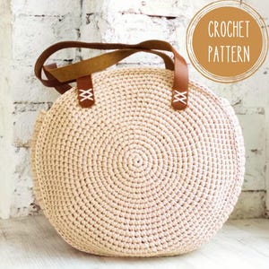 May include: A round, beige crochet bag with brown leather handles. The bag features a circular design and is adorned with brown leather straps attached with white stitching. The image also includes a circular graphic that says "CROCHET PATTERN".