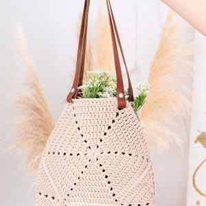 Op de afbeelding: Een lichtroze gehaakte tote bag met bruine leren hengsels. De tas heeft een cirkelvormig ontwerp met opengewerkte details. De tas wordt vastgehouden door een hand.