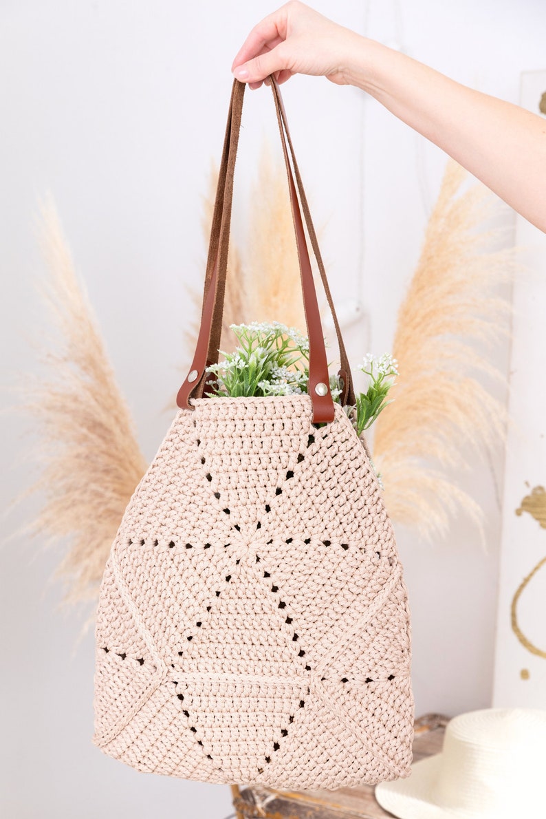 Op de afbeelding: Een lichtroze gehaakte tote bag met bruine leren hengsels. De tas heeft een cirkelvormig ontwerp met opengewerkte details. De tas wordt vastgehouden door een hand.
