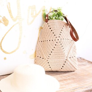 Op de afbeelding: Een beige gehaakte tote bag met een bruine leren band en een klein boeket groene planten erin. De tas ligt op een houten oppervlak naast een witte strohoed.