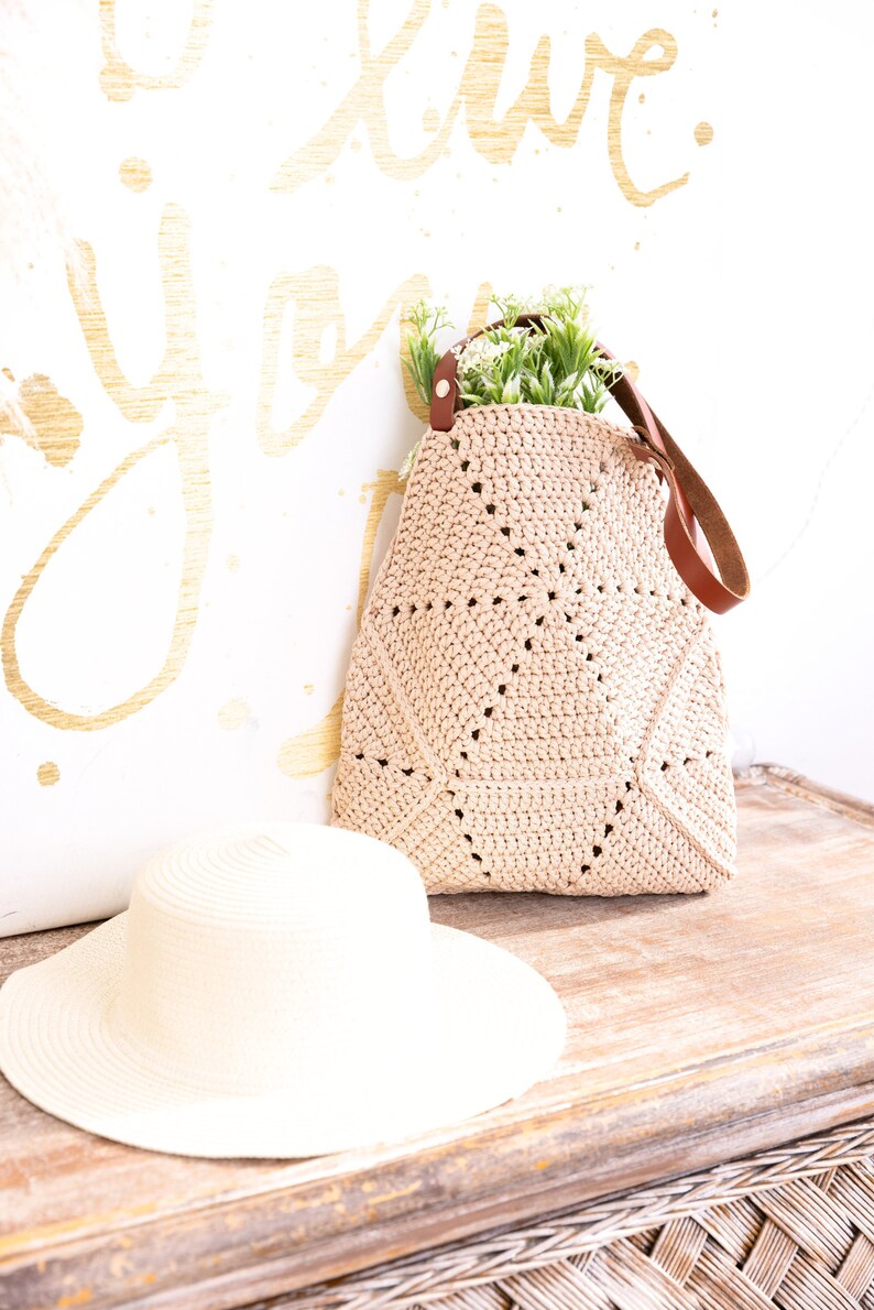 Op de afbeelding: Een beige gehaakte tote bag met een bruine leren band en een klein boeket groene planten erin. De tas ligt op een houten oppervlak naast een witte strohoed.