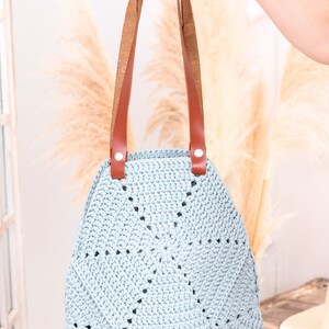 Op de afbeelding: Een lichtblauwe gehaakte tote bag met bruine leren hengsels en zilveren hardware. De tas heeft een cirkelvormig ontwerp met een stervormig patroon.