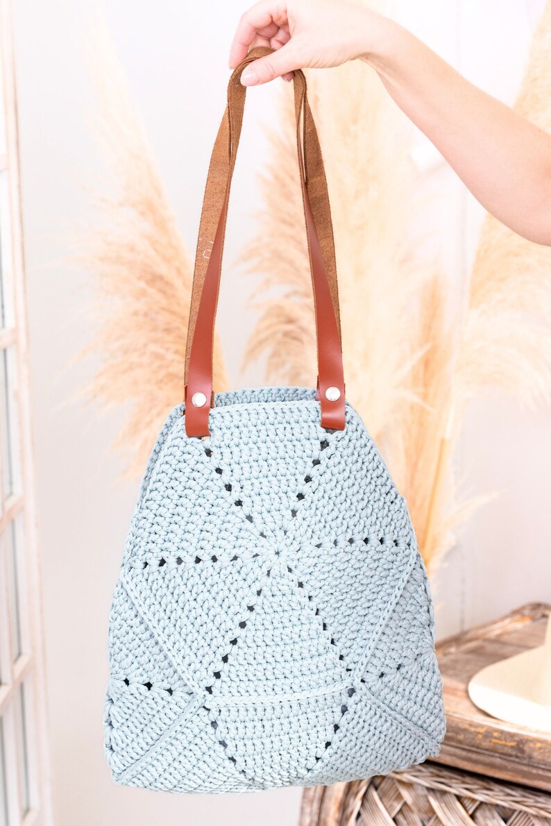 Op de afbeelding: Een lichtblauwe gehaakte tote bag met bruine leren hengsels en zilveren hardware. De tas heeft een cirkelvormig ontwerp met een stervormig patroon.