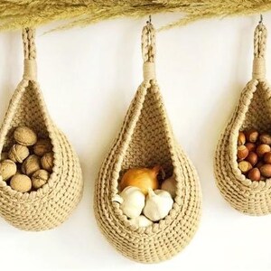 May include: Three beige, teardrop-shaped hanging baskets made of thick, woven cord. Each basket features a sturdy loop at the top for suspension. The baskets contain walnuts, garlic bulbs with a yellow onion, and hazelnuts, respectively.