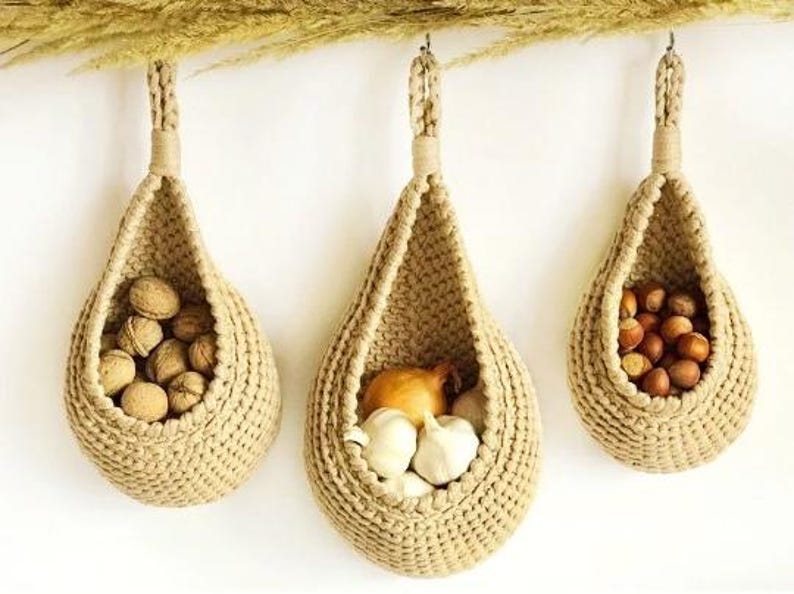 May include: Three beige, teardrop-shaped hanging baskets made of thick, woven cord. Each basket features a sturdy loop at the top for suspension. The baskets contain walnuts, garlic bulbs with a yellow onion, and hazelnuts, respectively.