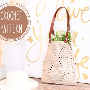 Op de afbeelding: Een gehaakte tote bag met bruine leren hengsels en een beige gehaakt lichaam. De tas staat op een houten oppervlak voor een wit canvas met de woorden "Love your life" geschreven in gouden verf.