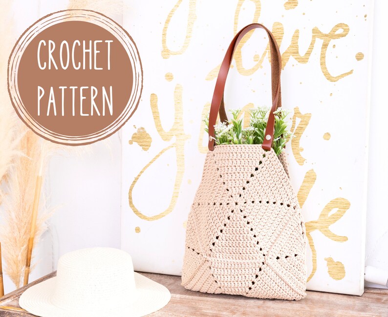 Op de afbeelding: Een gehaakte tote bag met bruine leren hengsels en een beige gehaakt lichaam. De tas staat op een houten oppervlak voor een wit canvas met de woorden "Love your life" geschreven in gouden verf.