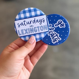 Peut inclure: Deux badges ronds à épingle. Le premier badge présente un motif vichy bleu et blanc avec une bannière horizontale blanche contenant le texte 'saturdays in LEXINGTON'. Le second badge est bleu uni avec des pois blancs et le texte 'Go Cats'.