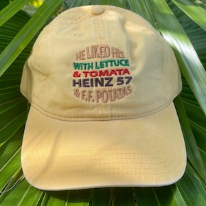 May include: A yellow baseball cap with the text "He liked his with lettuce & tomata Heinz 57 & f.f. potatas" embroidered in green, red, and black.