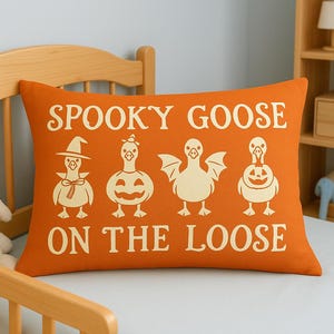 May include: Orange rectangular pillow with the text "Spooky Goose on the Loose" in white. The pillow features four cartoon geese in Halloween costumes. The geese are in a row, each with a unique design.