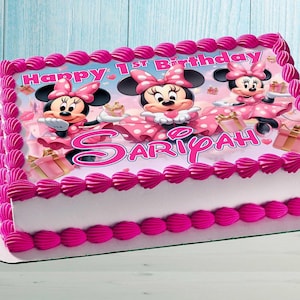 Op de afbeelding: Een rechthoekige verjaardagstaart versierd met roze glazuur en met Minnie Mouse. De taart heeft de woorden "Happy 1st Birthday" en de naam "SARIYAH" in het roze. Minnie Mouse is afgebeeld in een roze jurk met stippen.