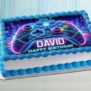 Puede incluir: Una tarta de cumpleaños rectangular con glaseado azul y una imagen decorativa de un mando de videojuegos. La imagen incluye el nombre "David" y las palabras "Happy Birthday" en texto blanco. La tarta está decorada con glaseado azul en los bordes.
