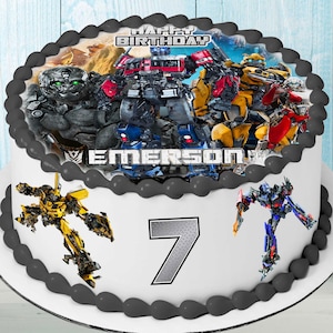 Pode incluir: Um bolo branco com uma imagem comestível com tema Transformers. A imagem apresenta Optimus Prime, Bumblebee e outros personagens dos Transformers. O bolo é decorado com uma borda de glacê preto e um número 7 prateado. O bolo é decorado com as palavras "Feliz aniversário Emerson".