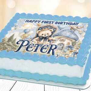 May include: A rectangular cake with blue frosting and a white base. The top features a blue and white image with a teddy bear wearing a crown, the name "Peter", and the text "Happy First Birthday".