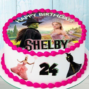 May include: A white cake with pink frosting and an edible image of the Wicked Witch of the West and Glinda the Good Witch from the film "Wicked". The cake is decorated with the name "SHELBY" and the number "24".