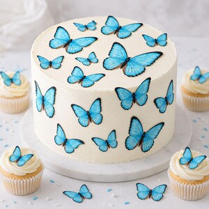 Puede incluir: Un pastel redondo con glaseado blanco liso decorado con múltiples mariposas azules, rodeado de cuatro cupcakes, cada uno con una mariposa azul encima, todo dispuesto sobre un soporte circular blanco.