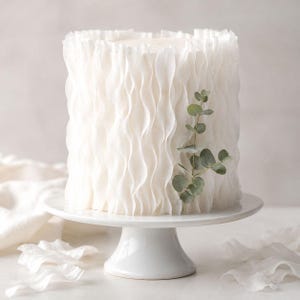 May include: A cylindrical white cake decorated with vertical ruffled layers of white icing. A single sprig of green eucalyptus leaves is placed against the side of the cake. The cake sits on a white pedestal stand with a smooth, rounded base.