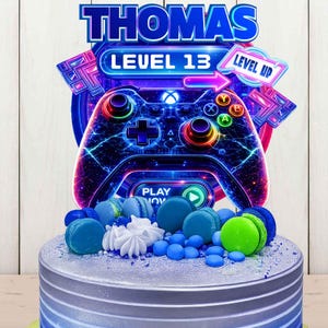 May include: A birthday cake with a silver frosting base, decorated with blue and green macarons, and white meringue. A topper features a video game controller design with the name "THOMAS" and the text "LEVEL 13" and "LEVEL UP".