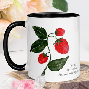 Peut inclure: Une tasse en céramique blanche avec un bord et une poignée noirs. La tasse présente une illustration aquarelle d'un plant de fraises avec des fraises rouges et des feuilles vertes. Le texte sur la tasse est "Je suis vieux. Je suis satisfait. Et tu étais mon but."