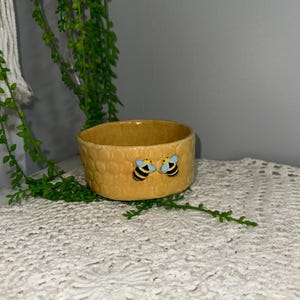 May include: A small yellow ceramic bowl with a honeycomb pattern and two bee figurines. The bowl is sitting on a white crocheted surface.