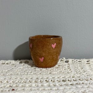 May include: A small, handmade brown ceramic cup with a textured surface. The cup is decorated with pink heart shapes. The cup is on a white, crocheted surface.